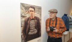 An elderly man wearing an orange plaid shirt and flat cap holds a camera, standing next to a large portrait of another man displayed on a museum or gallery wall.