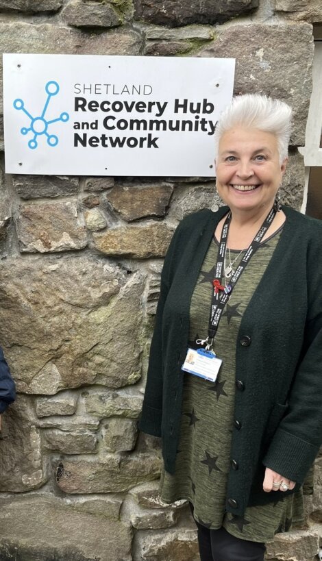 A person with short gray hair stands smiling in front of a stone wall beside a sign reading "Shetland Recovery Hub and Community Network." They are wearing a green dress, black cardigan, and lanyards.