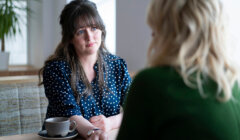 Two women are sitting at a table in a café, engaged in a conversation. One woman has dark hair and is wearing a polka-dot blouse, while the other has blonde hair and is wearing a green sweater.