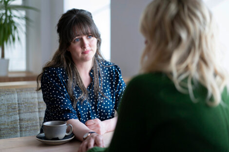 Two women are sitting at a table in a café, engaged in a conversation. One woman has dark hair and is wearing a polka-dot blouse, while the other has blonde hair and is wearing a green sweater.