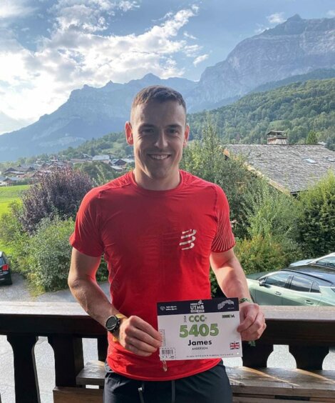 A man in a red shirt holds up a race bib with the number 5405, standing on a balcony with a mountain view in the background.