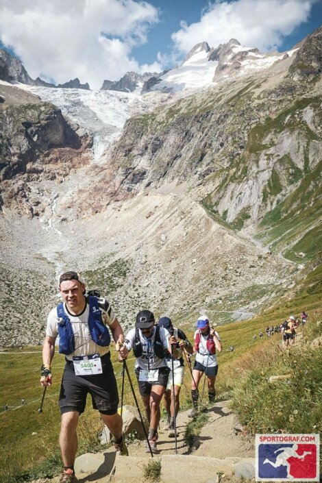 A group of runners with trekking poles ascend a steep mountain trail surrounded by rocky, grassy terrain and snow-capped peaks under a partly cloudy sky.