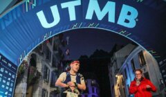 A runner crosses the finish line under the UTMB arch at night, wearing a headband and bib number 5405. Another person in red at the side appears to be clapping. Buildings are visible in the background.