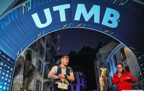 A runner crosses the finish line under the UTMB arch at night, wearing a headband and bib number 5405. Another person in red at the side appears to be clapping. Buildings are visible in the background.