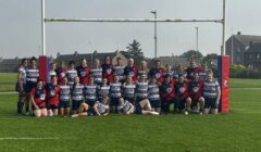 A group photo of two female rugby teams posing under a goalpost on a grassy field with housing in the background.