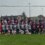 A group photo of two female rugby teams posing under a goalpost on a grassy field with housing in the background.