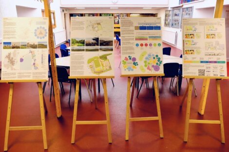 Four easels displaying large informational posters with maps, diagrams, and text are set up in a room with a red floor and chairs in the background.