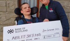 Two people holding a large cheque from the Bank of Scotland, made out to "Ability Shetland" for £1,000, dated 5th September 2024. The person on the right is wearing an "Abilities" branded jacket.