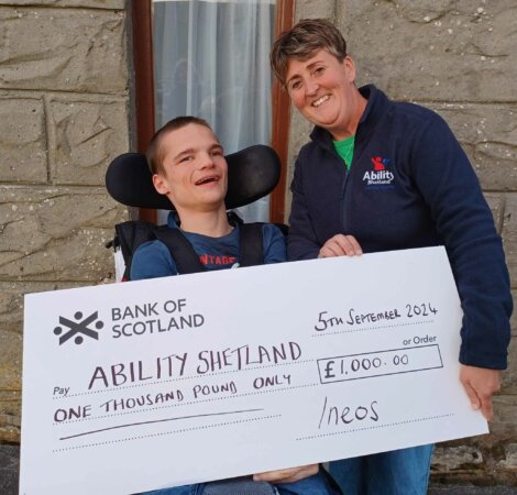 Two people holding a large cheque from the Bank of Scotland, made out to "Ability Shetland" for £1,000, dated 5th September 2024. The person on the right is wearing an "Abilities" branded jacket.