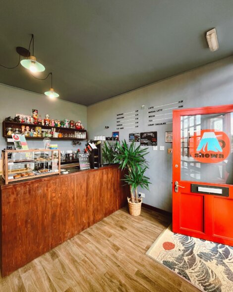 A small coffee shop interior with a wooden counter, assorted decorations, a potted plant, and a red door. A menu board is visible on the wall behind the counter.