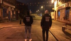 Two people wearing "Youth Work Team" jackets walk down a dimly lit, empty street at night. Buildings and colorful triangular flags are visible in the background.