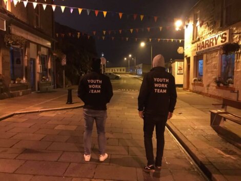 Two people wearing "Youth Work Team" jackets walk down a dimly lit, empty street at night. Buildings and colorful triangular flags are visible in the background.