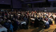 A large audience sits at tables in a dimly lit auditorium, attentively facing a stage not visible in the image.