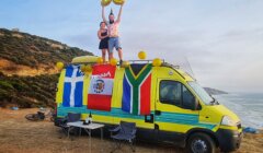 Two people on top of a yellow van holding balloons shaped like "30" with various flags on the van, two camping chairs and a table set up nearby, located on a coastal cliffside.