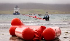 Large red buoy markers extend from the shore into the water toward a large vessel in the distance, with two smaller boats and several people working near the markers.