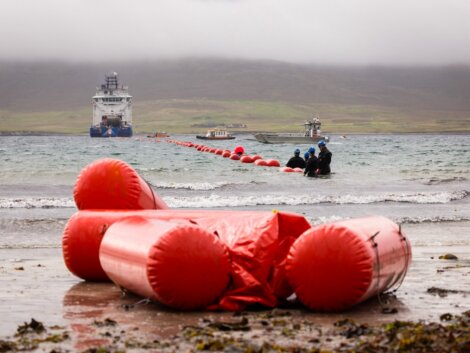 Large red buoy markers extend from the shore into the water toward a large vessel in the distance, with two smaller boats and several people working near the markers.