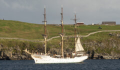 A tall ship with multiple masts and sails is anchored near a rocky shoreline with grassy hills and a few buildings in the background.