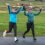 Two people in athletic clothing jog on a paved path, smiling and raising their arms exuberantly. A grassy field and distant buildings are in the background.