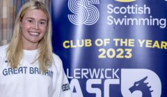 A person wearing a "Great Britain" shirt stands smiling next to a banner that says "Scottish Swimming Club of the Year 2023." The banner also features the Lerwick ASC logo.