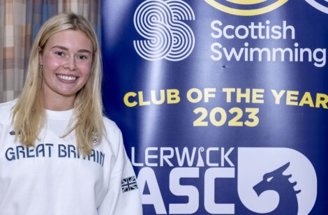 A person wearing a "Great Britain" shirt stands smiling next to a banner that says "Scottish Swimming Club of the Year 2023." The banner also features the Lerwick ASC logo.