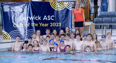 A group of children in swimwear pose in a pool next to a sign reading "Scottish Swimming" and "Lerwick ASC Club of the Year 2023." An adult in an orange shirt stands next to the group.