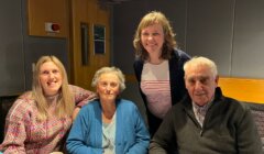 Four people sitting together at a table, with three smiling at the camera. The group consists of two older individuals and two younger women. They are indoors, in a room with grey walls and wooden accents.