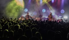 A band performs on stage with guitars, drums, and colorful lighting while a large crowd of fans watches and raises their hands.