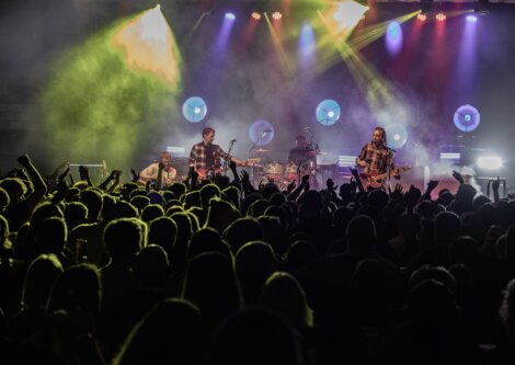 A band performs on stage with guitars, drums, and colorful lighting while a large crowd of fans watches and raises their hands.