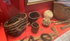 A display case showcases various woven baskets, balls of natural fibers, and bundles of raw materials. The items are arranged on a red surface.