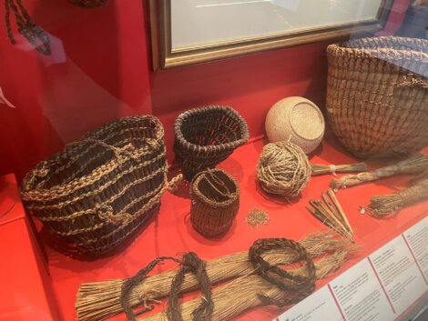 A display case showcases various woven baskets, balls of natural fibers, and bundles of raw materials. The items are arranged on a red surface.