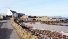 A narrow road winds past a white house and stone buildings by a rocky coastline with sparse vegetation and a small sandy beach. The sky is clear with light clouds.