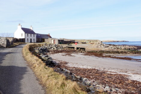 A narrow road winds past a white house and stone buildings by a rocky coastline with sparse vegetation and a small sandy beach. The sky is clear with light clouds.