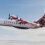 A Logan Air aircraft with tartan tail design is mid-flight against a partly cloudy sky. The plane's propellers are in motion.