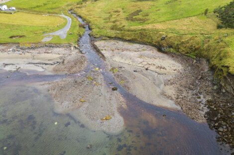 Aerial view of a small stream flowing into a beach inlet, surrounded by green grassy landscape and a gravel road.