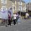 A group of people participate in a community march, carrying banners and balloons, walking down a residential street in daylight.