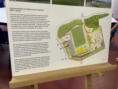 Display board showing a site plan with proposed improvements for pedestrian and vehicular access. The board is on an easel in an indoor setting with chairs in the background.