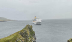 Large cruise ship sails through a misty sea near a rocky coastline.