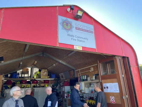 A red building with a sign that reads "Scottish Fire and Rescue Service, Walls Community Fire Station." People inside are engaged in conversation. Equipment and uniforms are visible in the background.