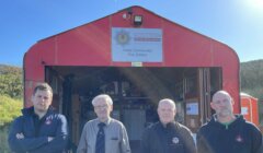 Four men stand in front of the Walls Community Fire Station, a red building with an open door. A sign above the entrance reads "Scottish Fire and Rescue Service.