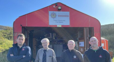 Four men stand in front of the Walls Community Fire Station, a red building with an open door. A sign above the entrance reads "Scottish Fire and Rescue Service.