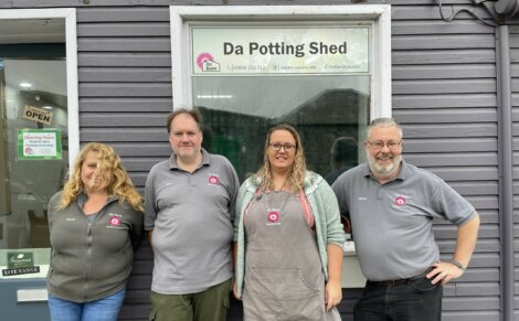 Four people standing in front of a grey building with a sign that reads "Da Potting Shed." They all wear grey shirts with matching logos.