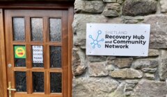 A wooden door next to a stone wall with a sign that reads "Shetland Recovery Hub and Community Network". The door has a smaller sign stating "IF THE DOOR IS LOCKED PLEASE KNOCK!.