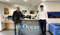 Two men stand at a reception desk with "Ocean Kinetics" signage. One man wears a plaid shirt and jeans; the other is in a white shirt and trousers. An aquarium and seating area are in the background.