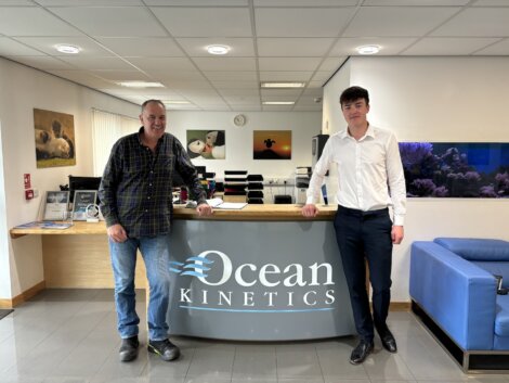 Two men stand at a reception desk with "Ocean Kinetics" signage. One man wears a plaid shirt and jeans; the other is in a white shirt and trousers. An aquarium and seating area are in the background.