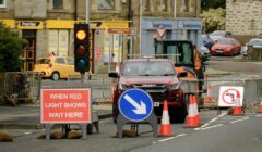 A red Isuzu truck stops at a road construction site with traffic cones, barricades, and signs including "When Red Light Shows Wait Here" and "No Left Turn" in front of a pharmacy and convenience store.