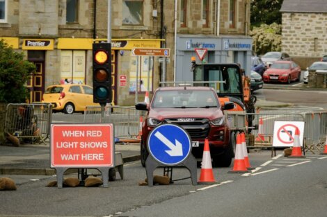 A red Isuzu truck stops at a road construction site with traffic cones, barricades, and signs including "When Red Light Shows Wait Here" and "No Left Turn" in front of a pharmacy and convenience store.