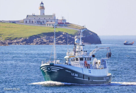 A green and white fishing boat sails near a rocky coastline with a lighthouse and buildings on a hill in the background. Another boat is visible further away in the water.