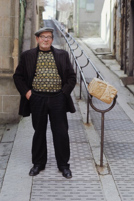 An elderly man stands on a narrow sidewalk with one hand in his pocket, wearing a beret, glasses, and patterned sweater. A wrapped package rests on a metal railing beside him.