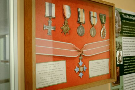 A wooden display case containing six military medals and two descriptive cards on a red background. The medals vary in design and color, with ribbons attached to some. Text and images are in the background.