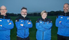 Four individuals standing outside with arms crossed, wearing blue and black jackets with team logos, and smiling for the camera. A grassy field is in the background.